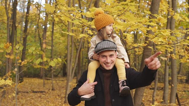 Father shows park in the autumn to daughter, holding her on shoulders, they are smiling, sunny day.