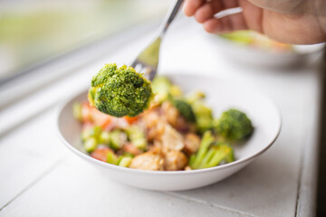 Fork picking broccoli from a broccoli and chicken salad next to a window