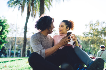 Young multiracial couple using smartphone and having fun on lawn in park