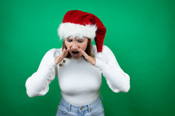 Young beautiful woman wearing a Santa hat and white sweater over green background shouting and screaming loud down with hands on mouth