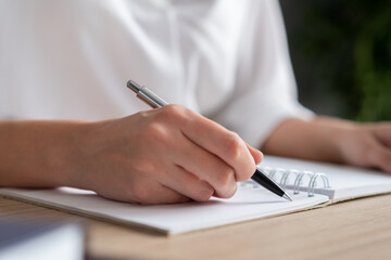 Female hand holding a pen making notes in notebook, closeup of blank paper list, woman office manager writing. Woman writing in office notebook, no face. Concept of writing