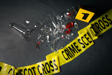 Broken bottle with blood, yellow tape and evidence marker on black slate background, flat lay....