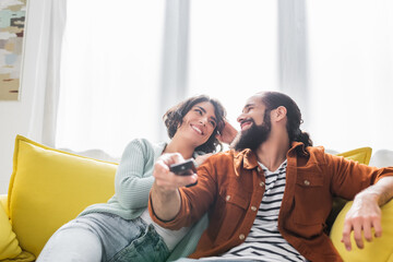 happy hispanic husband and wife looking at each other while watching tv at home