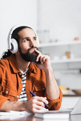 thoughtful hispanic man holding hand near face while listening audio lesson at home