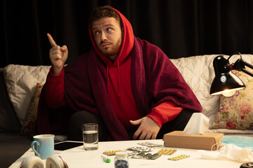 Pills, drugs. Man wrapped in a plaid trying to protect himself from somebody's sick near by, looks disguasted, angry, sad sitting on sofa at home indoors. Healthcare and medicine, ill prevention.