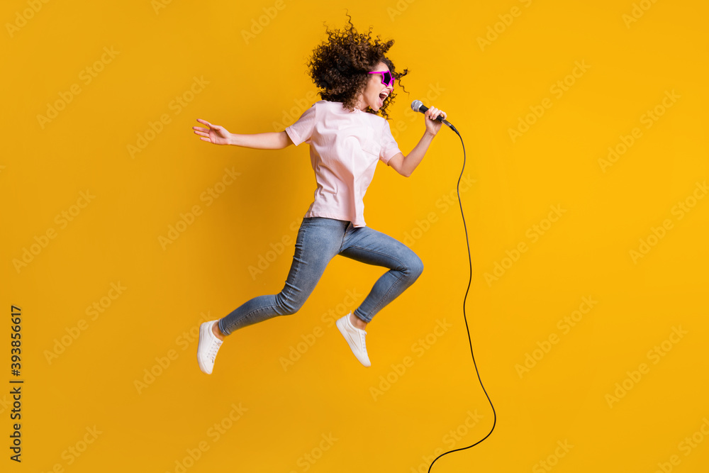 Wall mural photo portrait full body view profile of woman jumping up singing into mic isolated on vivid yellow 