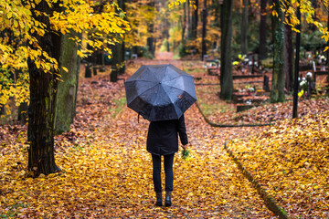 Woman with umbrella walking in cemetery at autumn. Lonely mourner in graveyard