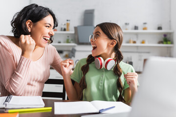 Excited hispanic mother and daughter with yes gesture looking at each other during online studying in kitchen on blurred foreground
