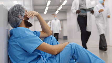 Upset and tired surgeon sitting on floor in hospital corridor - obrazy, fototapety, plakaty