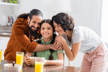 Hispanic parents hugging smiling daughter during breakfast on blurred foreground