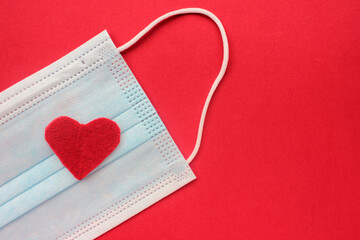 Disposable covid face mask with red heart on a red background. Safe Valentine`s day concept. Top view, copy space