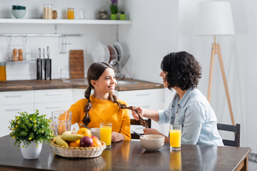 Hispanic woman touching hair of daughter near breakfast in kitchen