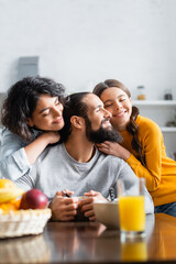 Smiling hispanic daughter and woman hugging father near breakfast on blurred foreground