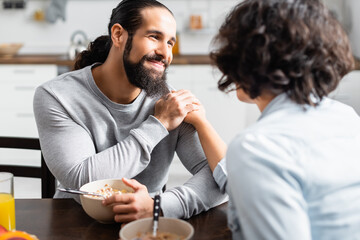 Smiling hispanic man holding hand of wife during breakfast on blurred foreground in kitchen