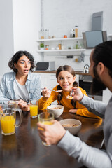 Excited hispanic woman looking at husband near daughter during breakfast on blurred foreground