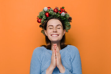 Cheerful woman in Christmas wreath hands folded in prayer beg about something good, dressed in...