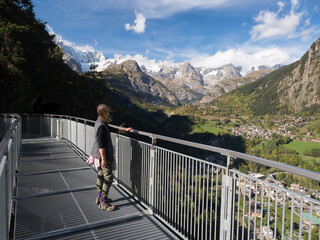 Fototapeta na wymiar A female walker stands on a high metal walkway looking at a dramatic view over a valley towards the Mont Blanc mountain range.Italy.