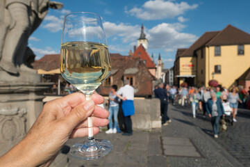 A glass of clear white wine is held up toasting the German city of Wurzburg. Deliberately blurred background of tourists walking and the buildings. Inverted reflection of the city skyline can be seen 