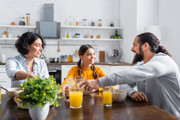 Smiling hispanic daughter looking at father near cereals and orange juice on table