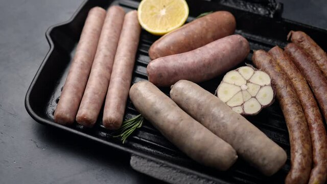The camera pans along a grill pan with meat sausages, sausages, lemon, rosemary and garlic. Place for text, dark background. 4K
