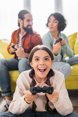 Fototapeta premium KYIV, UKRAINE - NOVEMBER 16, 2020: excited hispanic girl playing video game near parents holding joysticks on blurred background