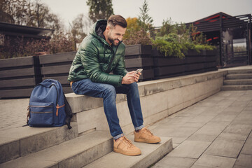 Profile side photo of attractive beaming young guy dressed green coat schoolbag having rest sitting stairs outdoors city street