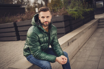 Photo of good mood attractive young guy dressed green coat having rest sitting stair outdoors city street