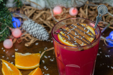 A glass of warming drink with a slice of orange, cinnamon and chocolate with coconut. Close-up. With a pine cone in the background.