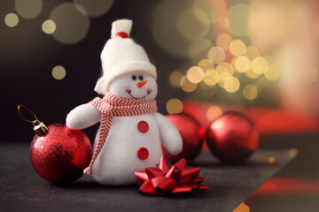 Closeup view of snowman toy, Christmas ball and bow on black table against blurred festive lights. Space for text