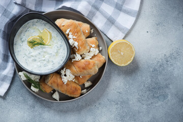 Greek mini phyllo rolls filled with feta and served with tzatziki dip, above view on a light-blue stone background with space