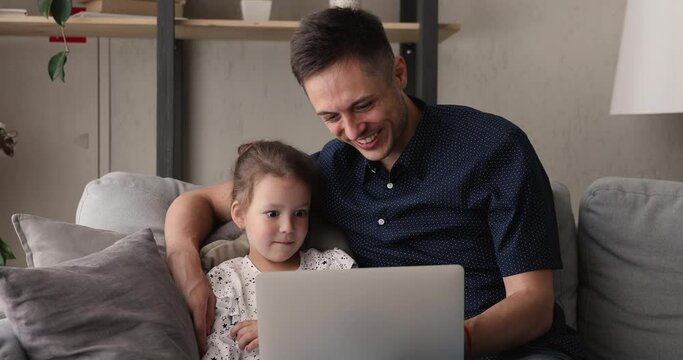 Father and 4s daughter relaxing on couch with laptop watching funny family movie, education program or cartoons together seated on soft comfy couch leaned on cushions. Weekend leisure pursuit concept
