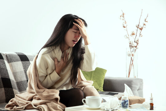 Sore Throat. Young Woman Wrapped In A Plaid Looks Sick, Ill, Sneezing And Coughing Sitting On Sofa At Home Indoors. Healthcare And Medicine, Ill Prevention, Seasonal Illness Symptoms And Self