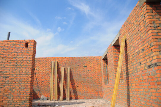 An inside view from an incomplete brick house construction with built walls and the blue sky instead of a roof. A house construction site with unfinished second floor and wooden rafters, eaves ready.