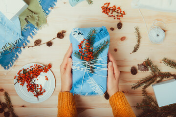 Zero waste, plastic free Christmas. Female hands holding gift box wrapped in fabric with spruce branch, berries on wooden background with natural materials. Flatlay. Sustainable holidays. Copy space.