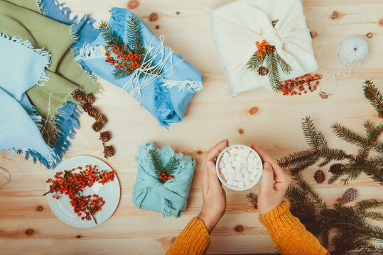 Female hands holding cup of cococa with marshmellow and just packed gift boxes wrapped in fabric and decorated with spruce branch, berries lying on wooden background. Zero waste, Sustainable Christmas