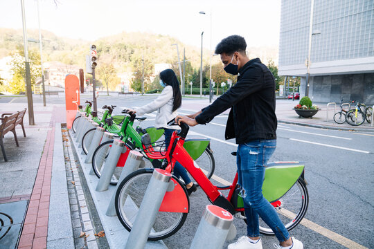A young Moroccan man and a Caucasian girl are picking up a rented electric bicycle in the street bicycle parking lot and wearing a face mask for the 2020 covid19 coronavirus pandemic. - Powered by Adobe
