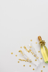 Festive white background with gold decoration , bottle of sparkling wine with two crystal glasses, shiny golden serpentine confetti and glittering snowflakes, copy space, top view