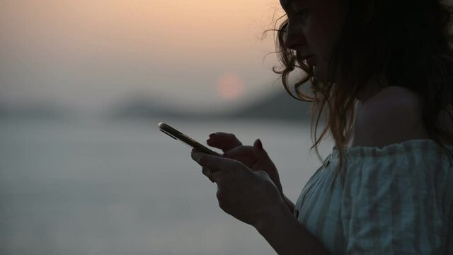 Young woman taping on a smartphone screen against sunset sky