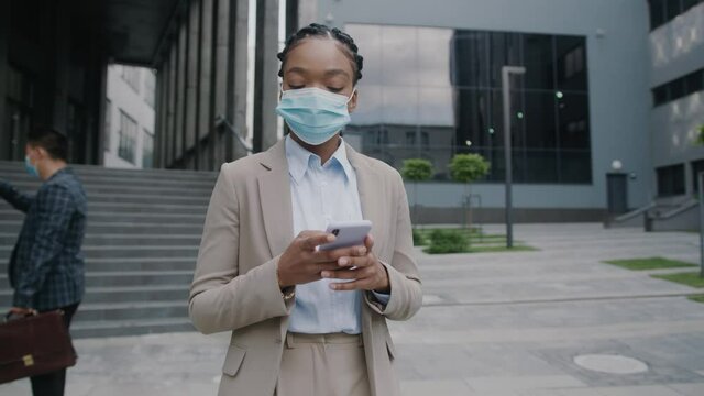 African American Female Office Manager With Protective Respiratory Mask Walking The Street Using Smartphone. Corporate People Communicating. Social Distance. Coronavirus.