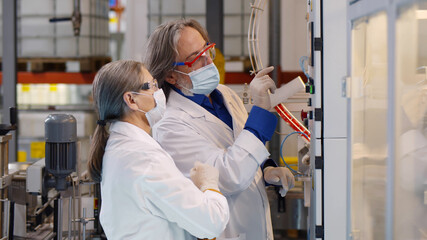 Mature male and female engineers in safety mask and white coat standing near operating machine