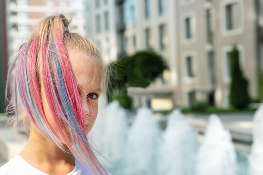Cute adorable caucasian little kid girl with multicolored pastel chalk pained dyed blond hair on city street with fountain background. Female child portrait pink colorful hairstyle smiling outdoors