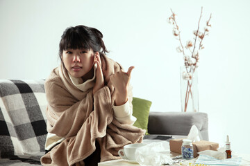 Obraz premium Pointing. Young woman wrapped in a plaid looks sick, ill, sneezing and coughing sitting on sofa at home indoors. Healthcare and medicine, ill prevention, seasonal illness symptoms and self-protection.