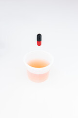 Small transparent plastic medicine drinking cup with red and black medicament capsule above it isolated on white