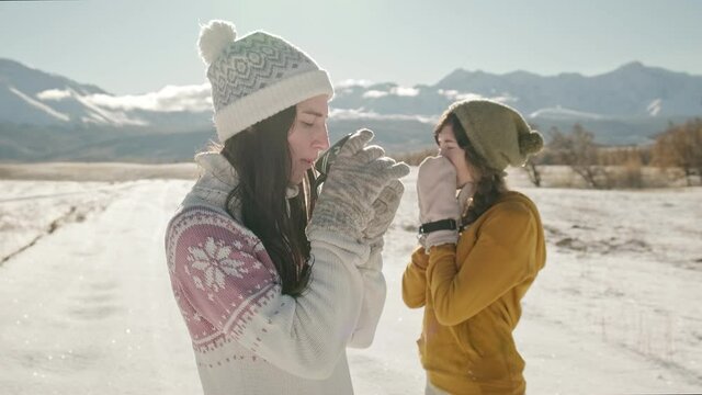 A young woman in a cozy sweater drinks a hot beverage from a mug, then shares it with her friend. Two female friends doing to explore snowy mountain landscape