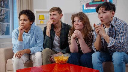 Multiracial friends sitting on couch eating chips and watching sports game on tv