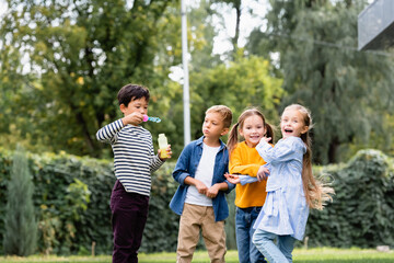 Fototapeta premium Cheerful multiethnic kids with soap bubbles standing in park