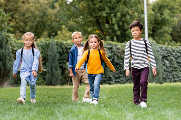 Obraz premium Multicultural schoolkids walking with backpacks on lawn in park