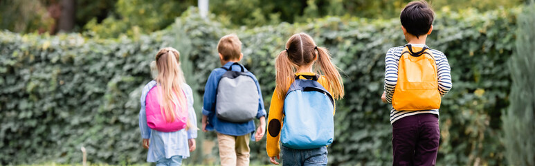 Back view of pupils with backpacks walking near friends on blurred background outdoors, banner