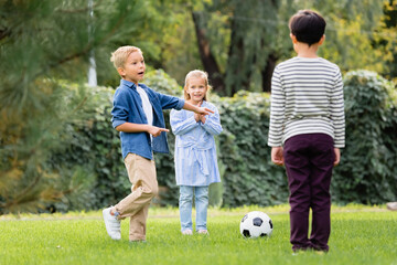 Excited boy pointing with fingers near football and friends in park