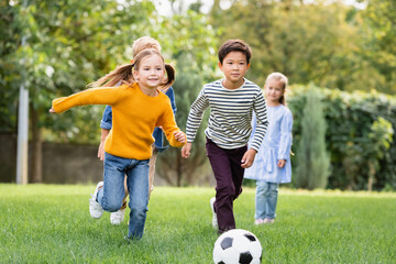 Cheerful multiethnic kids playing football near friends on blurred background in park